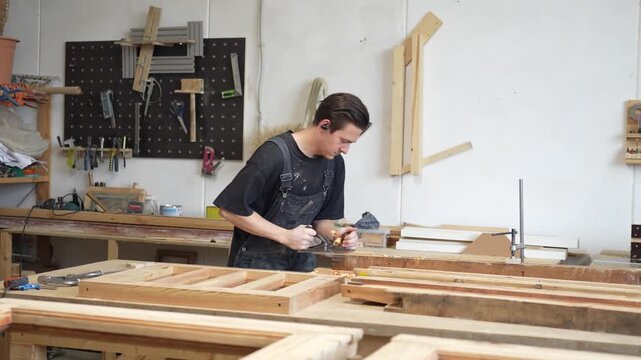 Concentrated man restorer hardworking in woodwork workshop on wooden window frame restoration, focused craftsman flattening and smoothing board surface with plane, handmade craft in carpentry.