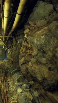 A narrow underground tunnel shows rocky walls and metal pipes. The area is lit by a dim light at the far end, creating shadows in the dark space. The ground is uneven with visible tracks.
