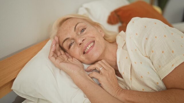 Woman lying on bed, hand to head and fingers near mouth, ring visible on finger, pillow and wooden headboard, pajama top with small dots in studio; serenity.