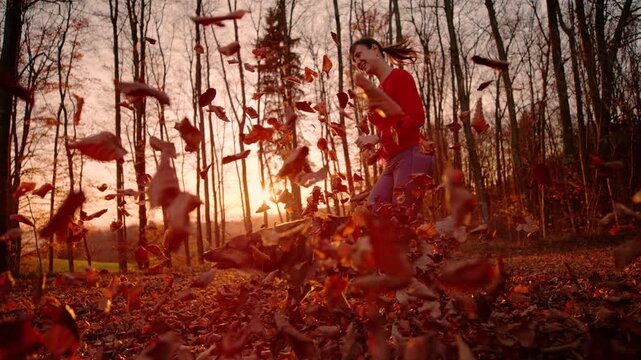 LENS FLARE, LOW ANGLE VIEW, SLOW MOTION: Smiling young woman runs with her dog along forest trail covered in dry autumn leaves. Flying foliage, warm sunset light, outdoor activity and seasonal charm.