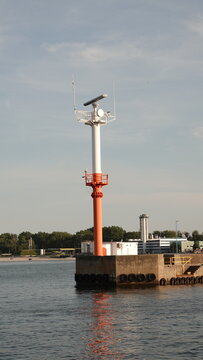 Zdjęcie przedstawia biało-czerwoną wieżę nawigacyjną z radarem, umieszczoną na betonowym falochronie u wejścia do portu. W tle widać spokojną wodę, linię brzegową z drzewami oraz pogodne niebo.