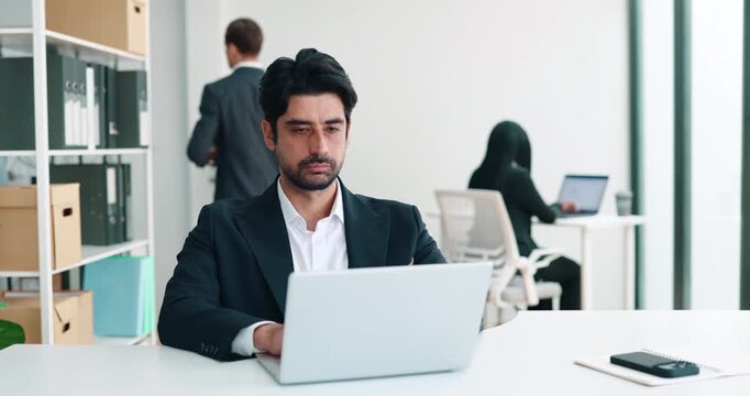 Portrait of Middle Eastern businessman faces laptop at desk as colleague walks in back. Concentrated man studies screen continuing office workday with focus