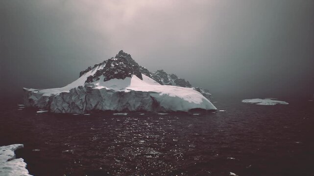 Close ice floe on black water, distant ice island and jagged peak under heavy clouds, light glints on ripples, scene evokes exploration and navigation risk,