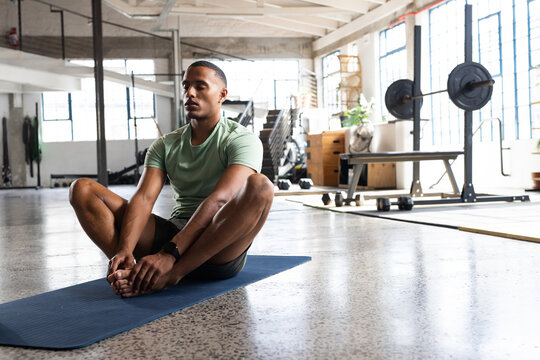 African American man sitting on blue mat in gym wearing green T-shirt and watch near dumbbells