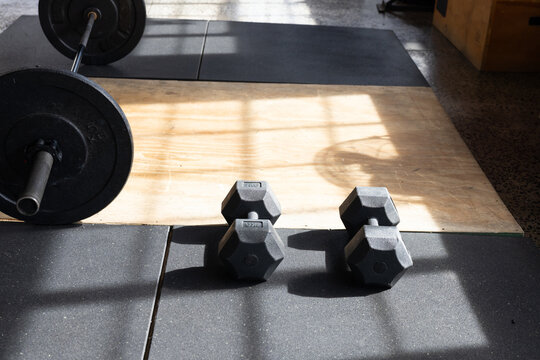 Black hex dumbbells are resting on rubber mats and casting window-grid shadows on platform in gym