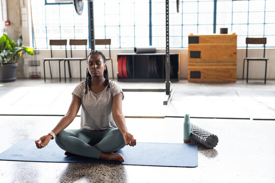 Adult woman sitting meditating on blue mat in studio, gray tee, teal leggings, foam roller beside