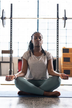 African American woman meditating on blue mat in gym with barbell, wearing gray tee, teal leggings