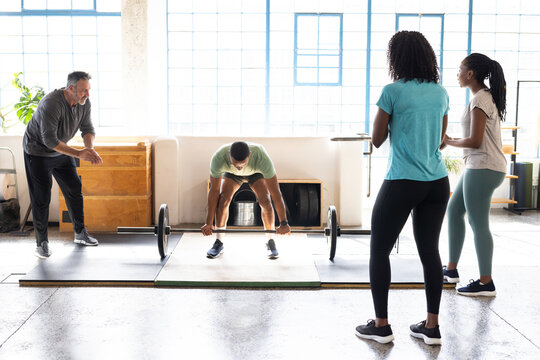 Diverse training group coaching and spotting male lifter preparing barbell on platform in gym
