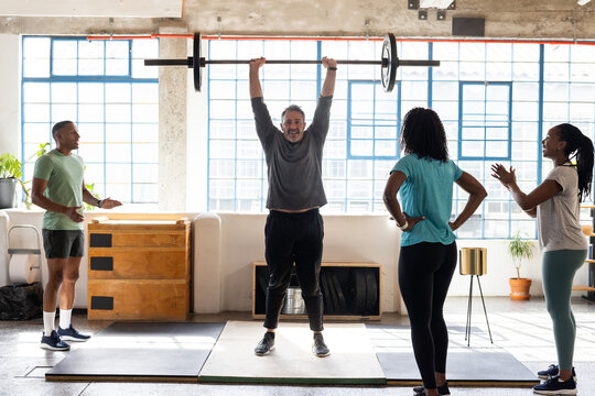 Diverse gym members surrounding man lifting barbell overhead on platform in bright loft gym