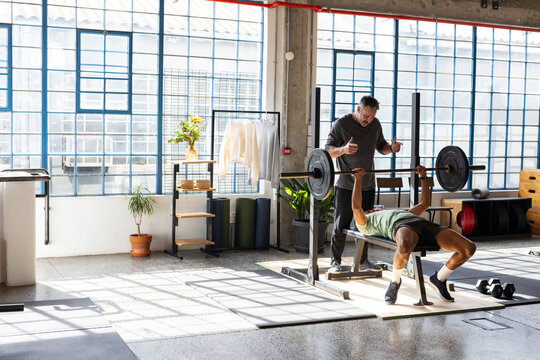 Male trainer spotting while male trainee pressing loaded barbell on bench at loft gym, copy space