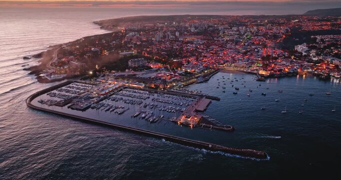 Cascais Marina and the Atlantic coastline in Portugal glowing with city lights at sunset, boats moored in the harbor with the vibrant town extending along the Portuguese shore. Aerial view panorama