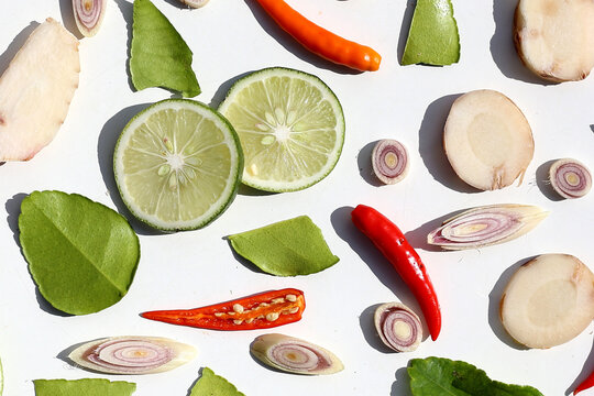 A colorful, close-up pattern of Thai herbs: lime slices, galangal, lemongrass, chilies, and kaffir lime leaves.