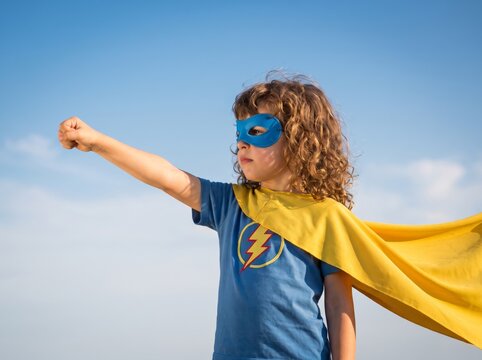 Brave young child dressed in a blue superhero costume with a yellow cape and mask stands proudly against a bright blue sky, extending a fist forward in a powerful, imaginative flying pose outdoors.
