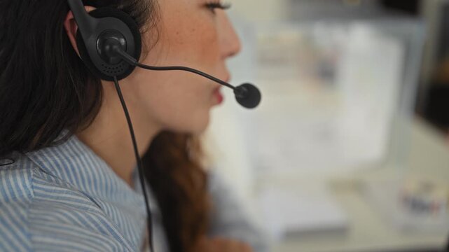Young hispanic woman wearing a headset leans forward and speaks into headset microphone with mouth visible at a desk in an office building; focus service.