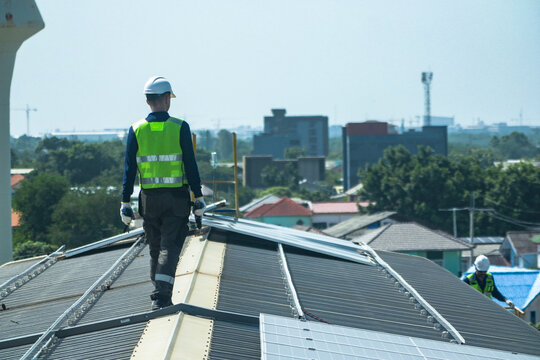 A renewable energy specialist in reflective workwear and helmet confidently pauses at the roof edge platform between inspections on a large corrugated factory rooftop under the tropical sun.