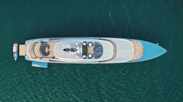Large luxury superyacht with tender boat moored at the stern, cruising on the blue green oceanwater, showcasing opulence and freedom in maritime travel and vacation. Top d own aerial view footage