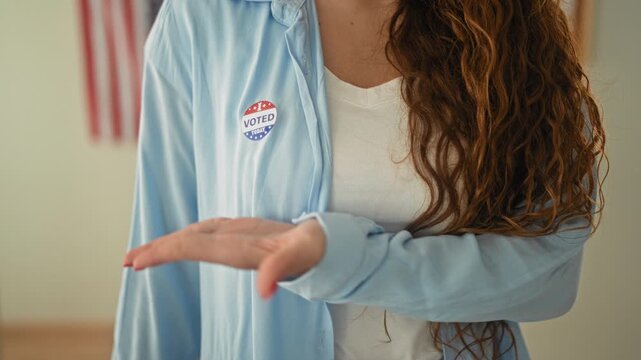 Woman points finger to voted sticker and shows open palm, long hair visible, american flag in frame in building; pride civic duty.