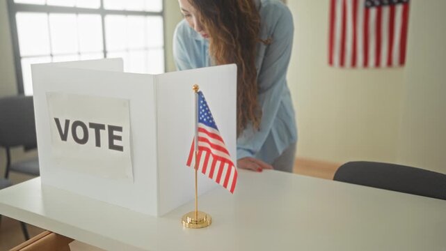 Young hispanic woman leans over voting booth with hand on table, scanning ballot beside small american flag in polling building; patriotic duty.