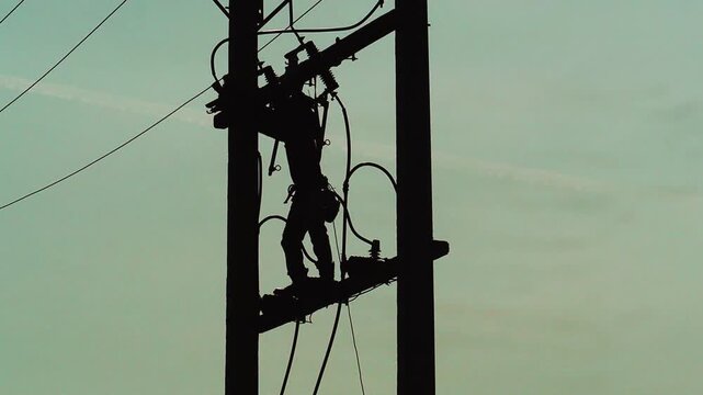 Silhouette shot of electrician lineman working high up on electric utility pole repairing power lines.