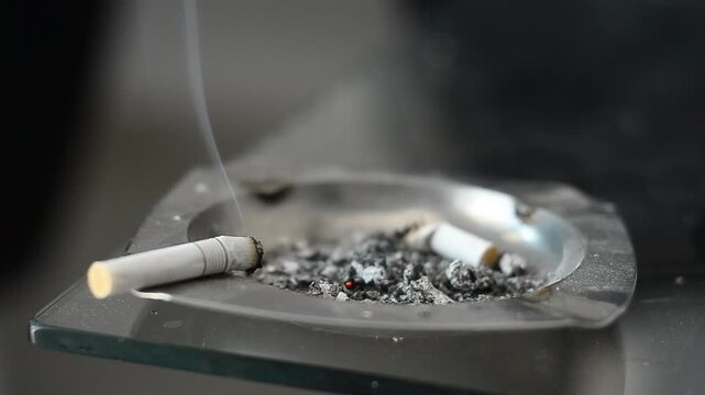 Hand drops cigarette into ashtray filled with smoke and ash