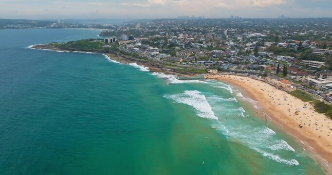 Manly Beach coastline extending towards the city skyline, featuring an ocean pool, golden sand filled with people relaxing and swimming in the blue waves on sunny summer day. Aerial view drone flight