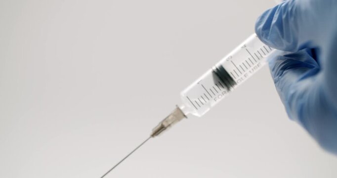 Medical professional's hand in a blue glove holding a syringe and pushing the plunger to eject the liquid, isolated on a clean white background. Medical treatment and vaccination concept