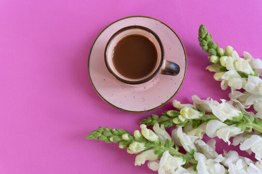 Pink table holds warm coffee cup. White flowers bloom beside the cup. Soft lighting enhances cozy feel. Perfect for morning or quiet moments.