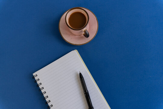 Blue surface holds coffee and notebook. Black pen rests beside open lined pages. Cup of coffee sits on matching saucer. Still, calm, quiet moment captured above. Ideal for work, study