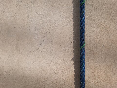 Blue braided rope hanging against a cracked beige wall with a sharp parallel shadow.