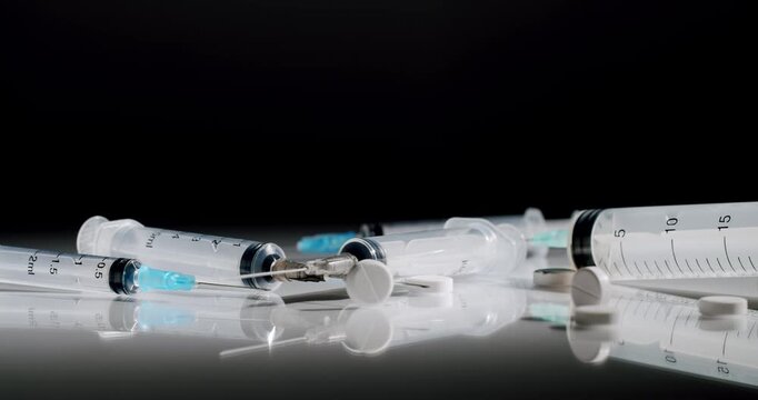 Medical syringes of various sizes and white pills lying on a reflective surface against a black background, representing concepts of healthcare, drug abuse, addiction, and vaccination