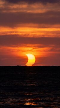 Partial solar eclipse at sunset over the sea with glowing orange sky and dark ocean waves creating a dramatic celestial event scene