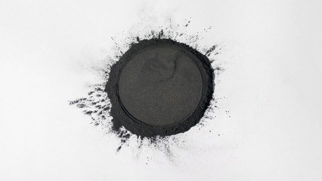 Shot following a vertical arc of a heap of black powder with a circular mark on a dim grey background, lit by orbiting light creating shifting shadows | Black cumin seed powder commercial concept