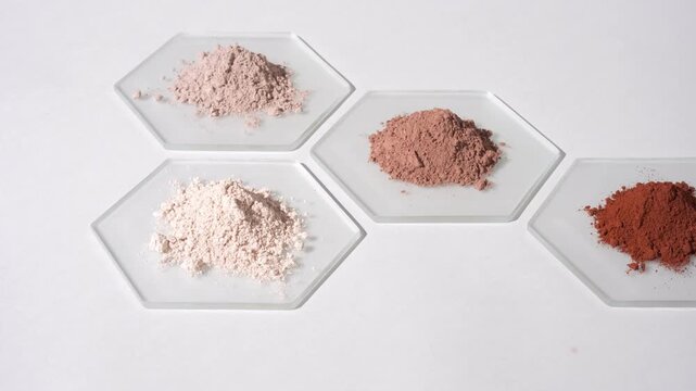 Vertical jib high-angle shot of piles of different color powders beige and brown on hexagonal watch glass pieces on light grey background lit by moving light casting shadows | Cosmetic ingredients
