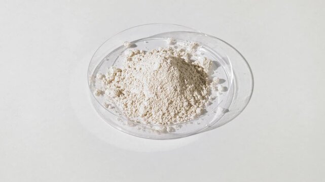 Vertical crane shot of white fine powder piled in a plastic petri dish on a light gray background illuminated by rotating light projecting shifting shadows | Dimethicone cosmetic ingredient concept