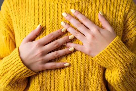 Close-up of female hands with pastel yellow nails wearing a matching yellow knitted sweater. Monochrome fashion aesthetic.