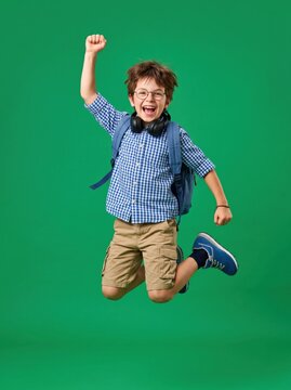 Happy energetic school boy jumping high with backpack and headphones on green background.