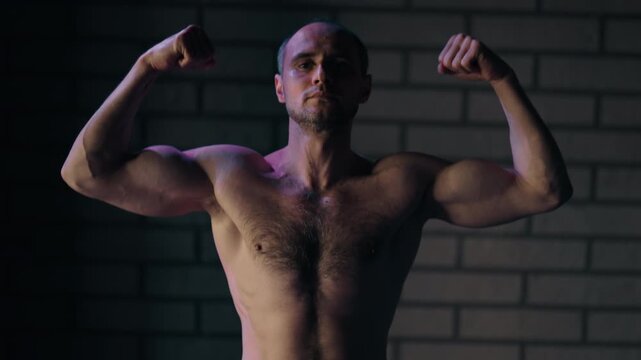 Athletic man flexing his muscles and showing his biceps in a dark gym after a workout. Strong bodybuilder posing for the camera with dramatic lighting, demonstrating his physical strength and power