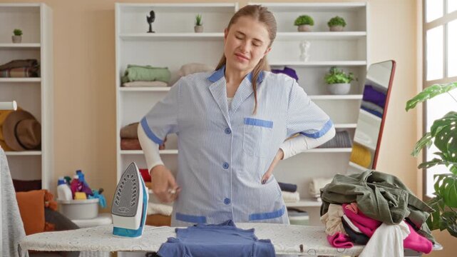 Woman cleaner in uniform ironing a shirt with iron on board, hand on lower back and face wincing while stacking clothes in a building; fatigue duty.