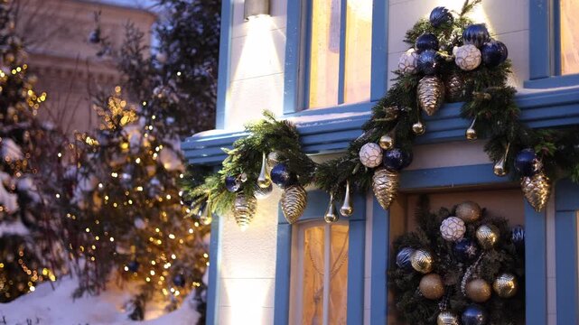 Cinematic Christmas exterior with a blue-trimmed house decorated with evergreen garlands, blue, white and silver ornaments, pinecones and warm lights. A snow-covered Christmas tree with glowing golden