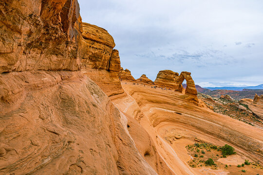 Sweeping view of the massive sloping sandstone bowl leading toward Delicate Arch, with tiny figures of hikers exploring the vast desert landscape.
