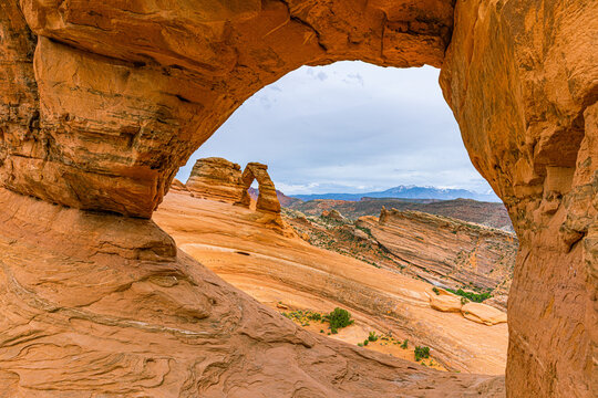 Iconic Delicate Arch standing on a sloping sandstone basin, beautifully framed through the rugged opening of a natural rock window in Arches National Park.