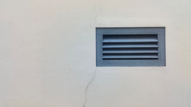 Close-up of a grey ventilation louver grille on a cracked white cement wall. Concept of architectural exterior detail, building utility design, and urban structure photography.