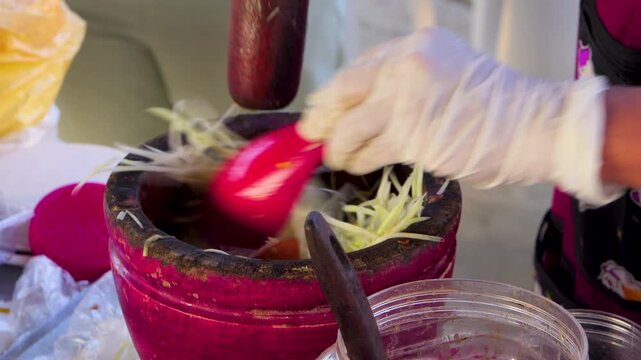 Street food vendor using red spoon and wooden pestle to mix authentic Thai papaya salad in mortar.