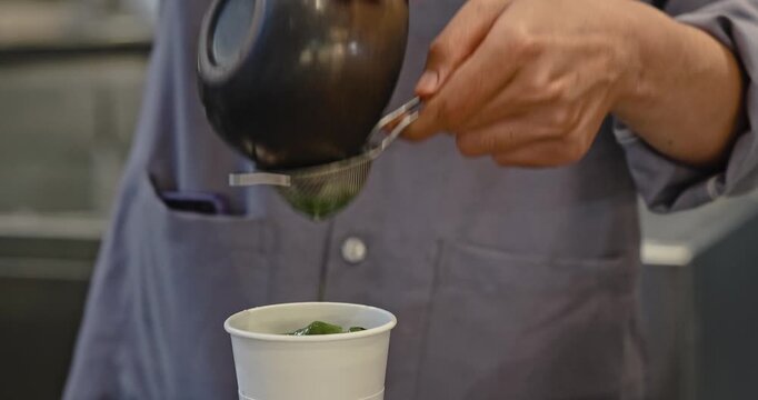 Barista hands pouring green matcha liquid through metal sieve into white paper cup, professional beverage preparation in cafe kitchen, culinary documentary style.