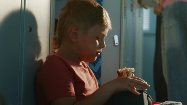 Schoolboy Sitting Comfortably on Floor Near Metal School Lockers, Eating Lunch From Food Container During Recess. Authentic Childhood, Education, School Routine, and School Lifestyle Concept.
