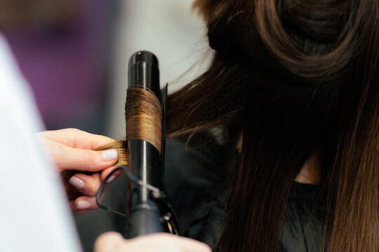 Professional Hairdresser Curling Client Long Brown Hair With Curling Iron. Hairstylist hands wrapping a strand of female hair around a hot curling rod in salon
