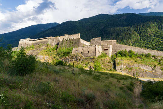 A massive stone fortress spans a steep hillside overlooking a forested mountain valley beneath a partly cloudy sky. Ancient defensive walls and towers rise above lush green grassy slopes below
