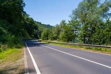 Fototapeta premium A smooth, curving asphalt road winds along a wooded hillside beside a distant river valley view on a sunny day. Dense green trees and a metal guardrail line the roadside under a bright blue sky