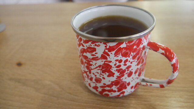 A hot cup of black coffee or tea served in a traditional red and white speckled enamel mug on a light wooden table.
