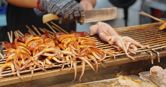 Squid skewers grilling on metal grill at Asian street food market, traditional seafood preparation, culinary travel and gastronomy documentary.